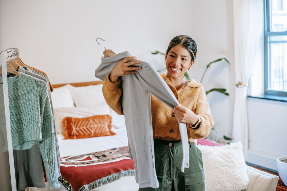 Cheerful ethnic female in trendy outfit considering shirt on hanger while standing near rack in bedroom