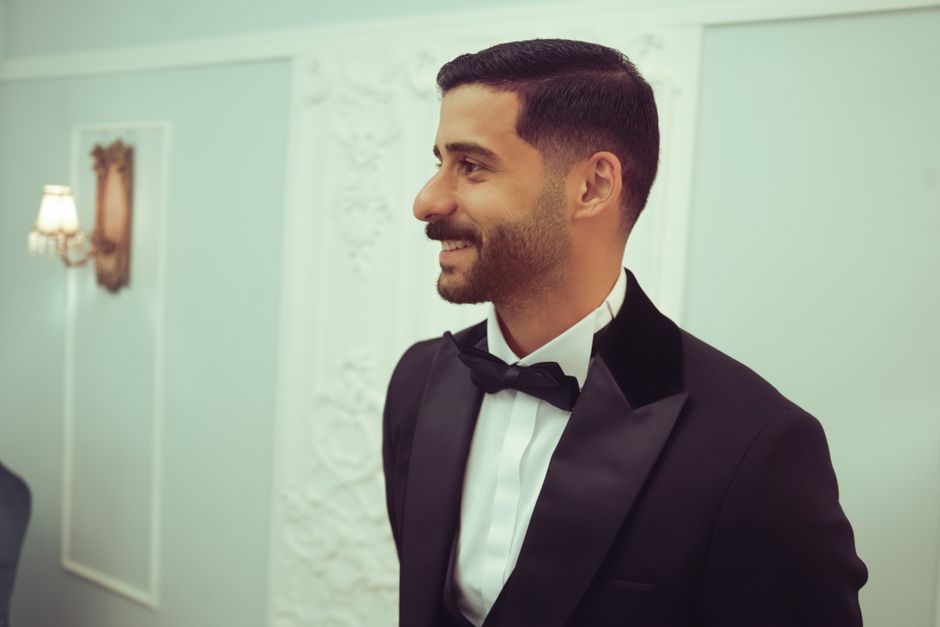 A handsome man in evening wear smiling indoors, exuding elegance and warmth.