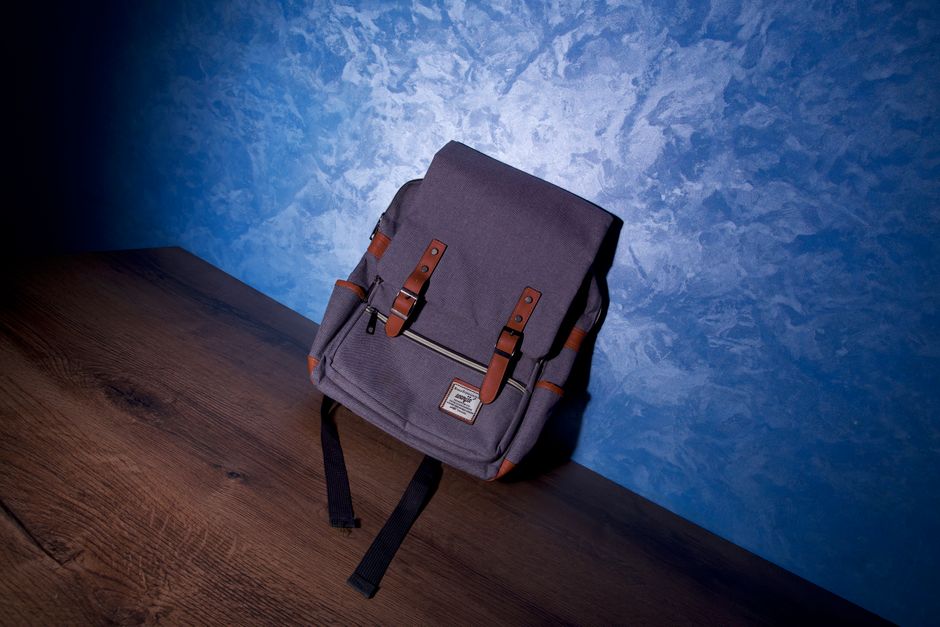 A modern backpack with leather straps sits on a wooden surface against a textured blue wall.