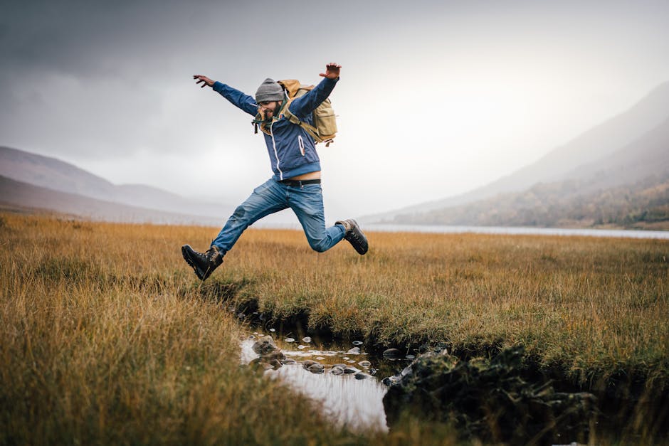A man jumps with excitement over a stream in a picturesque, open landscape.