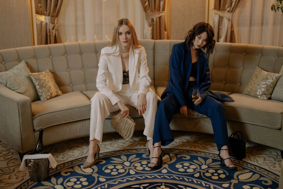 Two women in luxurious suits sit on a sofa holding champagne, exuding elegance and glamour.