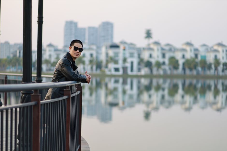 Man in sunglasses and leather jacket leans on railing near a city lake with modern buildings.