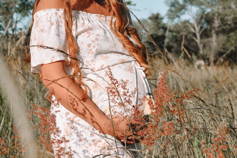 A serene portrait of a pregnant woman in a floral dress surrounded by sunlit grasses in a field.