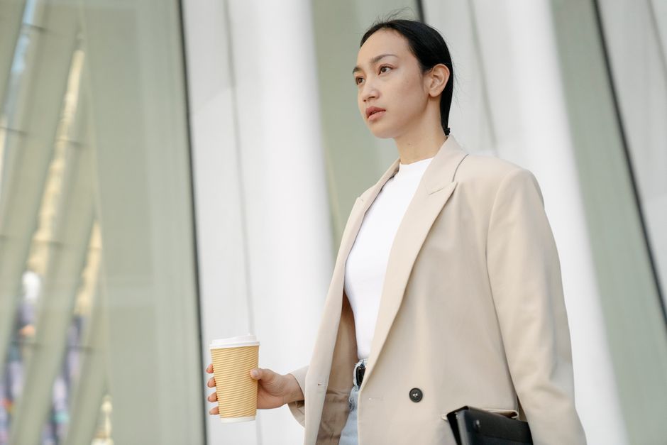 Confident Asian woman in beige coat holding coffee cup and tablet outdoors.