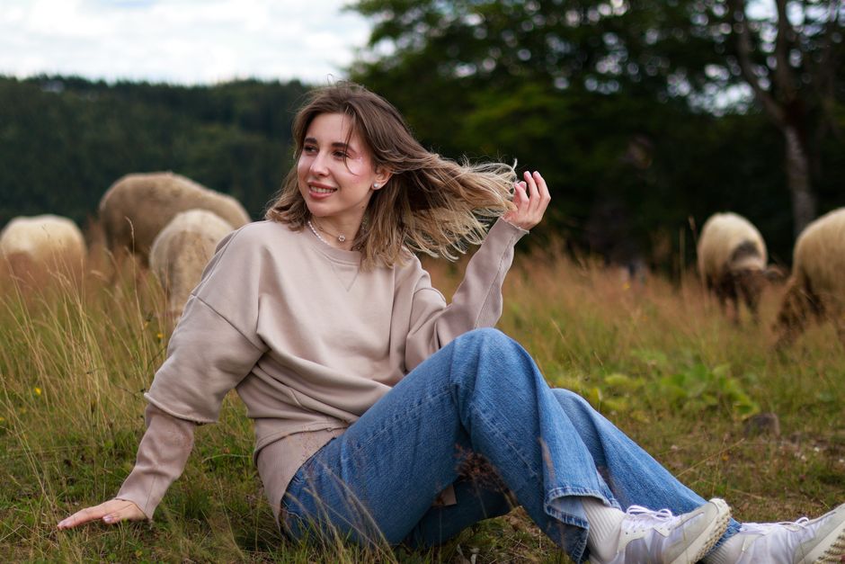 Young woman in a sweater and jeans sitting in a rural field with sheep, enjoying a peaceful moment outdoors.