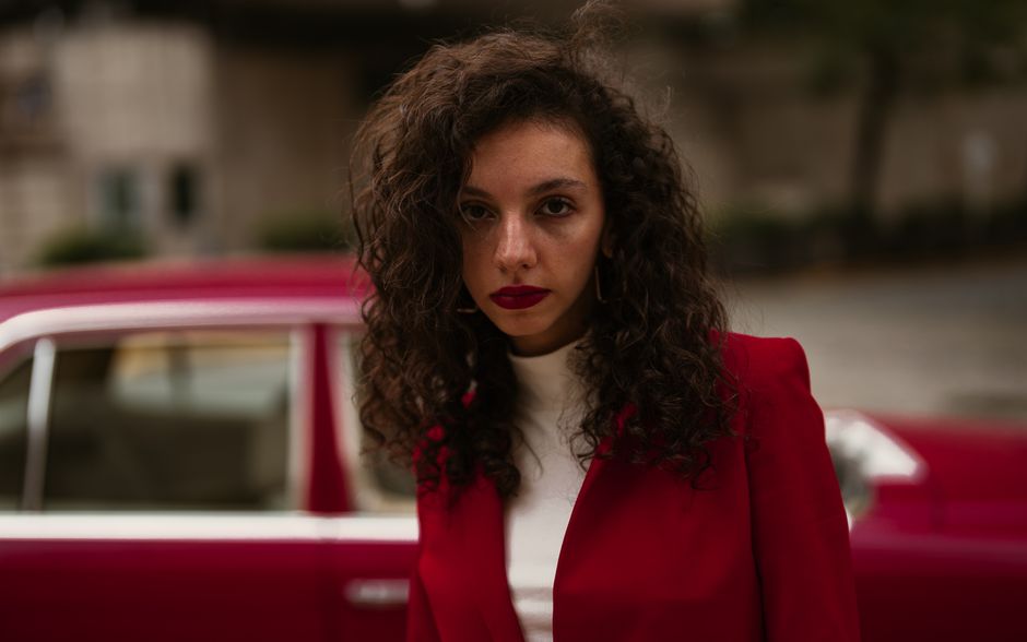 Fashionable woman in red blazer poses by a vintage car on a city street.