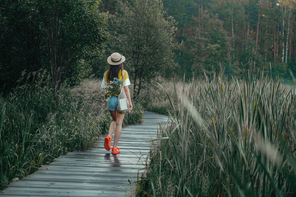 A woman in casual attire walks along a wooden pathway through lush greenery.