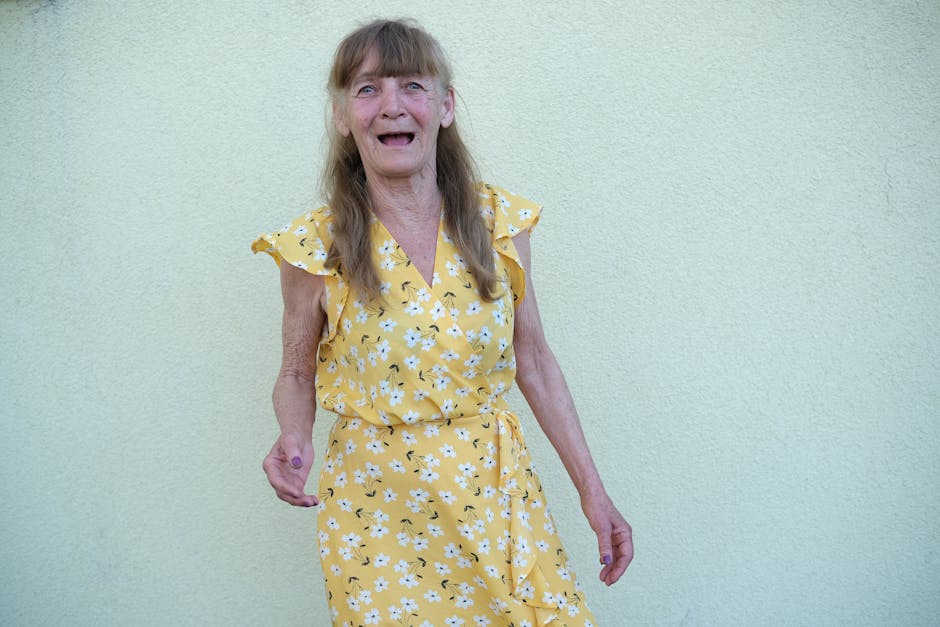 Joyful senior woman wearing a floral yellow dress, expressing happiness against a plain background.