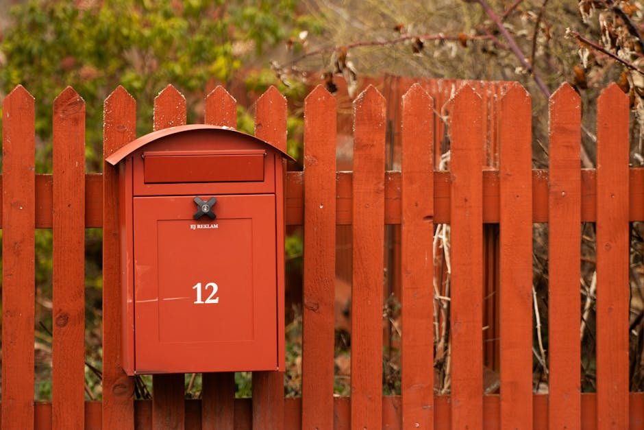A red mailbox mounted on a matching wooden fence surrounded by vibrant greenery in Jönköping, Sweden.