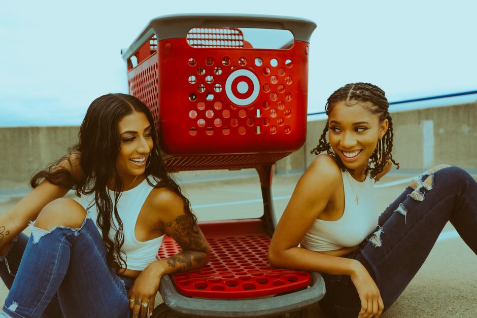 Two fashionable women sitting with a red shopping cart, sharing smiles in an outdoor setting.