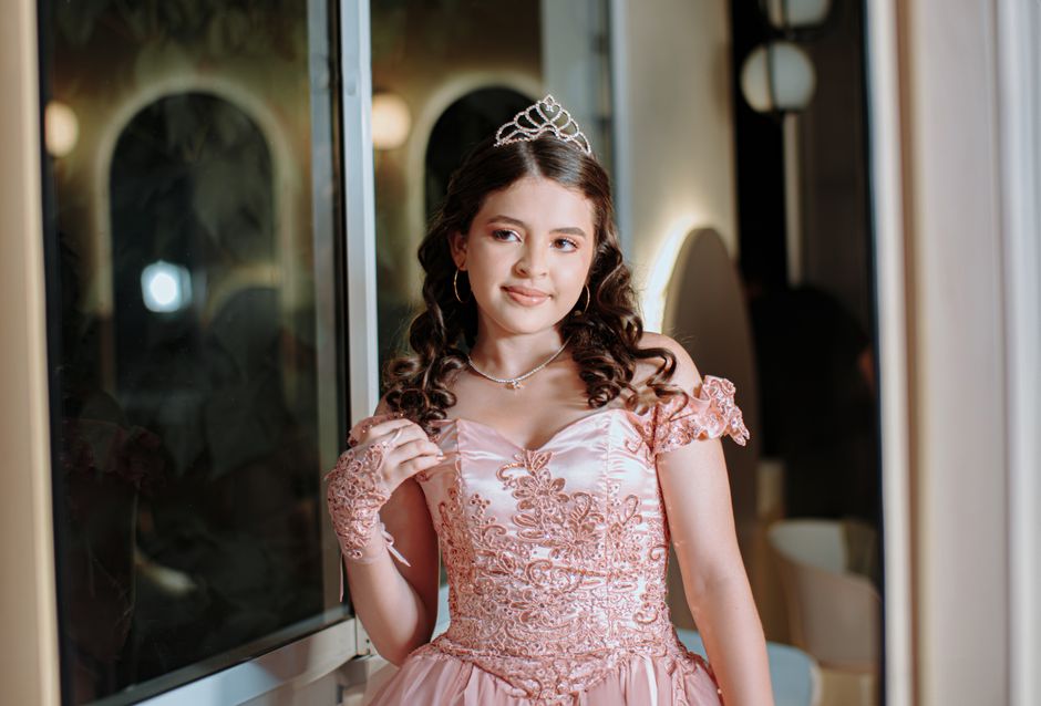 Charming young woman in a pink lace ballgown and tiara posing indoors by a window, radiating elegance and glamour.