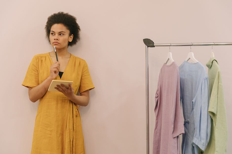 A woman in a yellow dress ponders creatively in a clothing studio with racks.