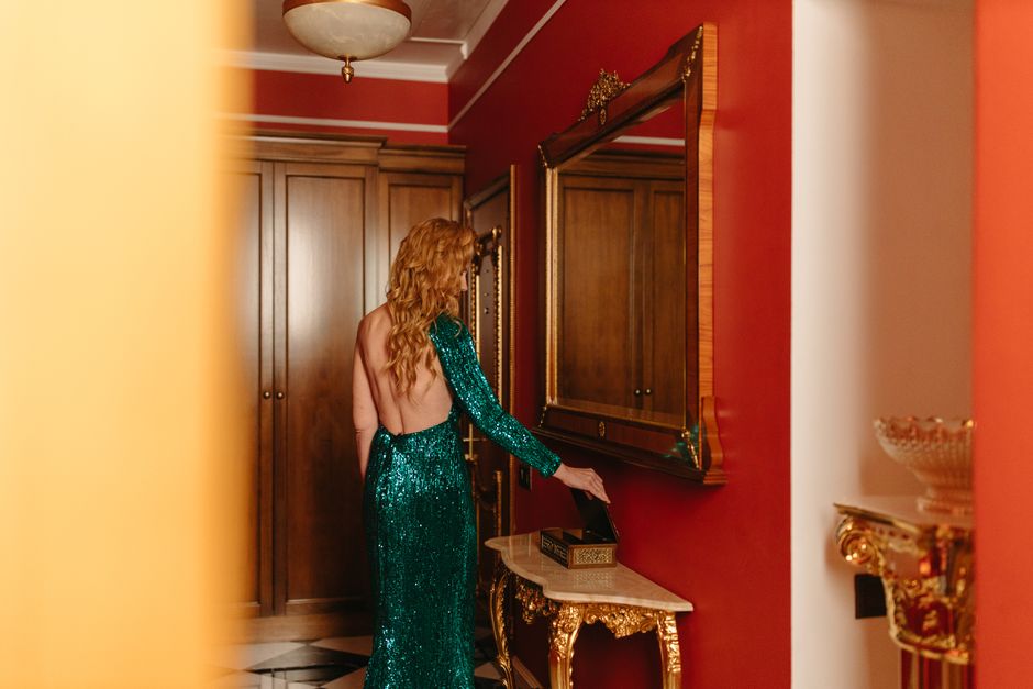 Woman in a sparkling dress admires herself in a mirror inside a luxurious room with rich decor.