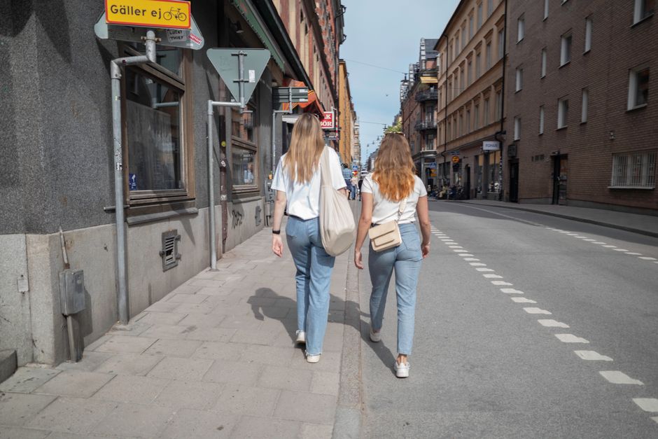 Two women with long hair walk down a city sidewalk on a clear day, showcasing urban life.