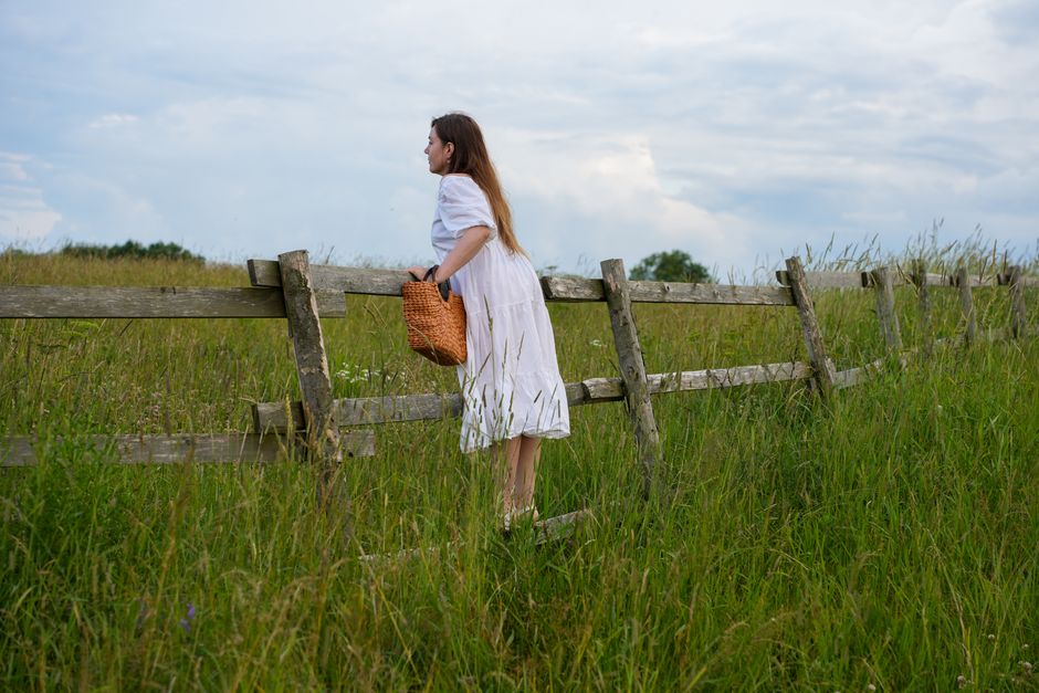 A woman in a white dress leans on a wooden fence in a serene rural field, carrying a basket.