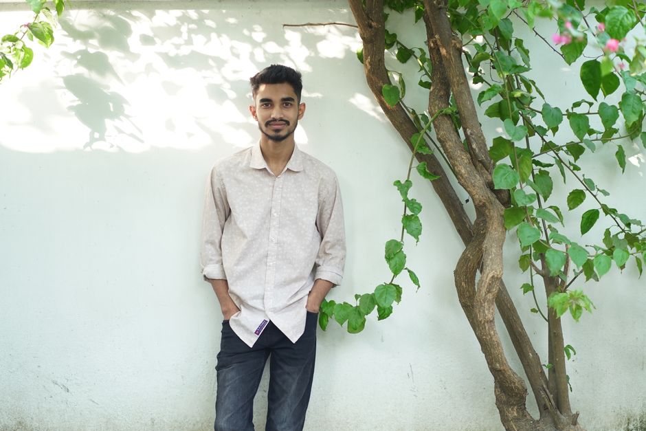 Casual young man leaning against a wall beside a tree in a sunny outdoor setting.