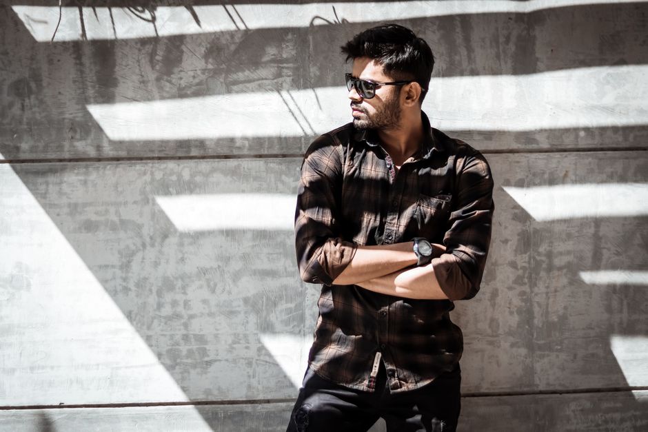 Man in stylish plaid shirt and sunglasses striking a pose against a gray concrete wall with dramatic shadows.