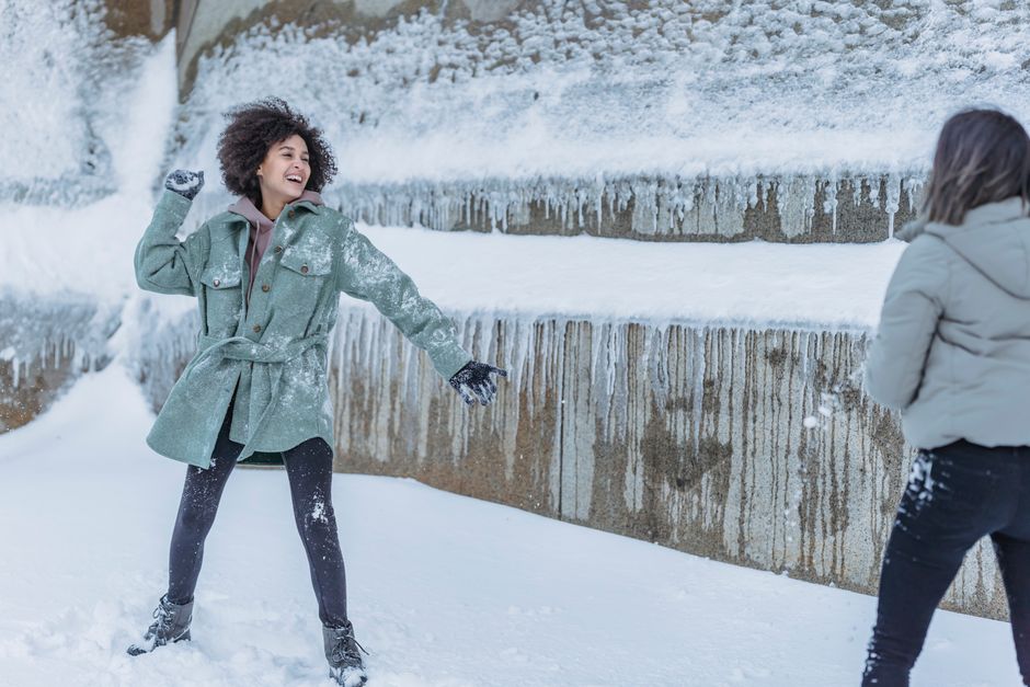 Laughing female friends in warm clothes standing near stone wall covered with snow and playing snowballs in winter