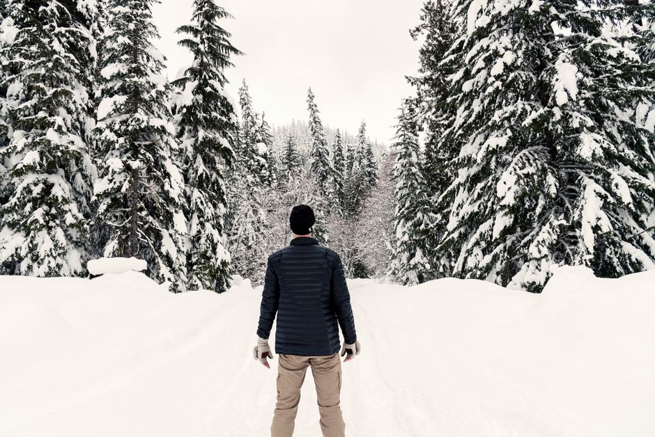 A man stands alone in a serene, snow-covered forest during winter, surrounded by tall evergreen trees.