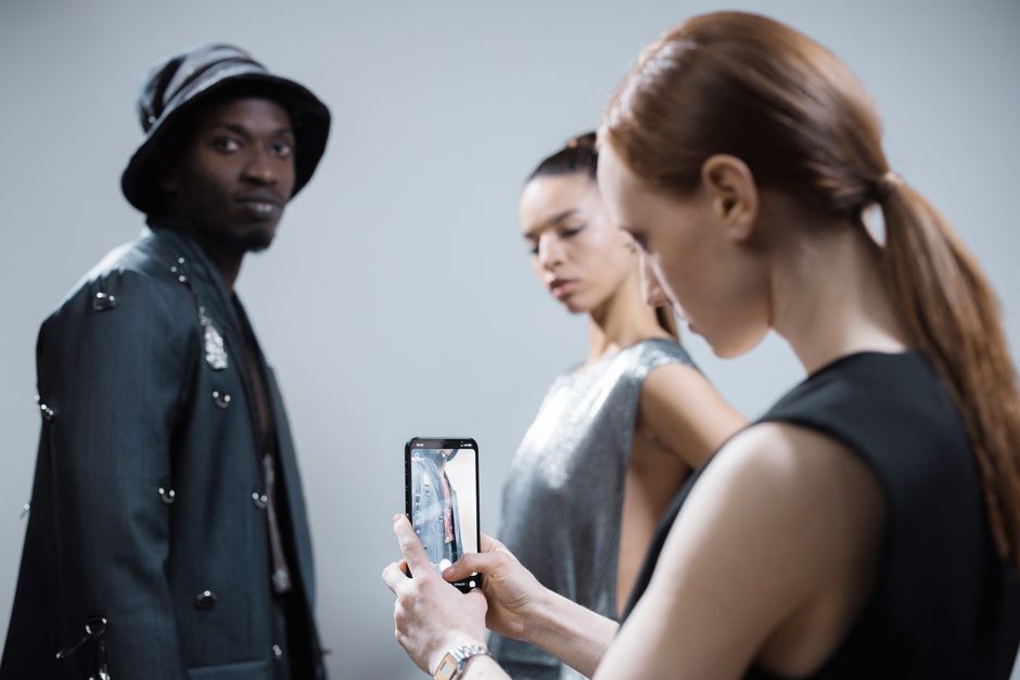 Model poses in studio with photographer capturing the scene on mobile.